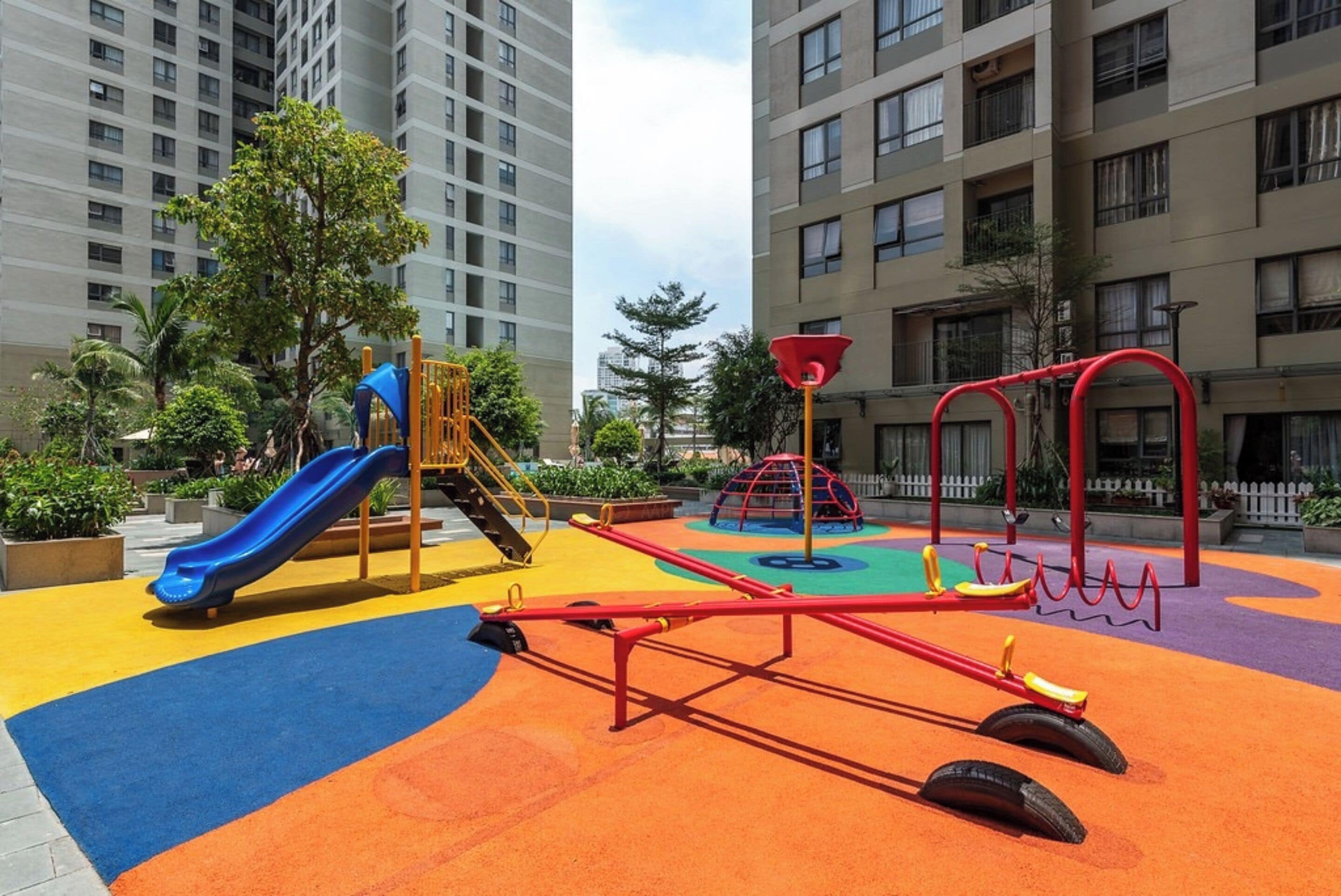 A playground with a slide, seesaw, and climbing structures, surrounded by apartment buildings.