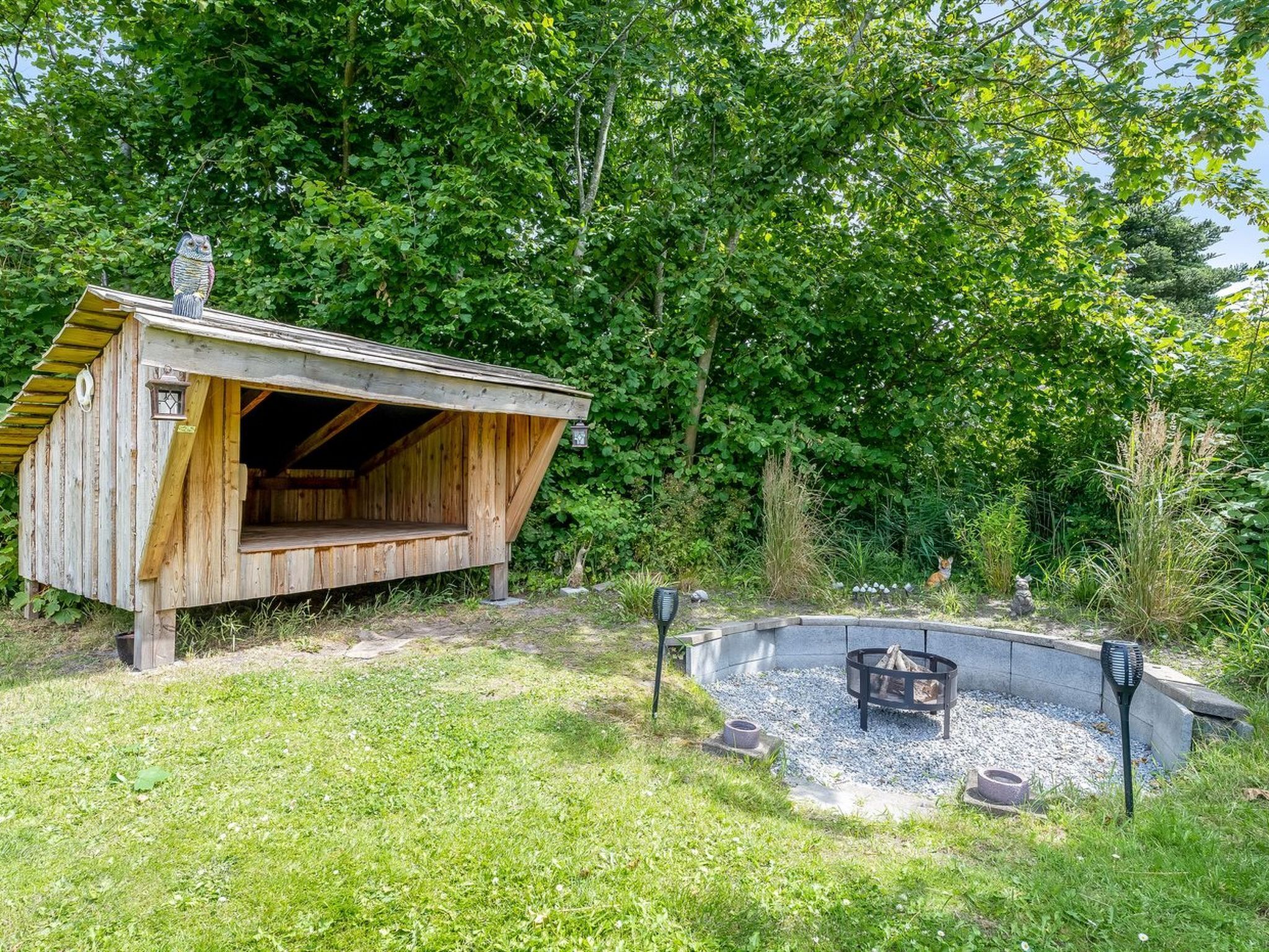 A wooden cabin with an open front, surrounded by a garden with a fire pit and stone lanterns.