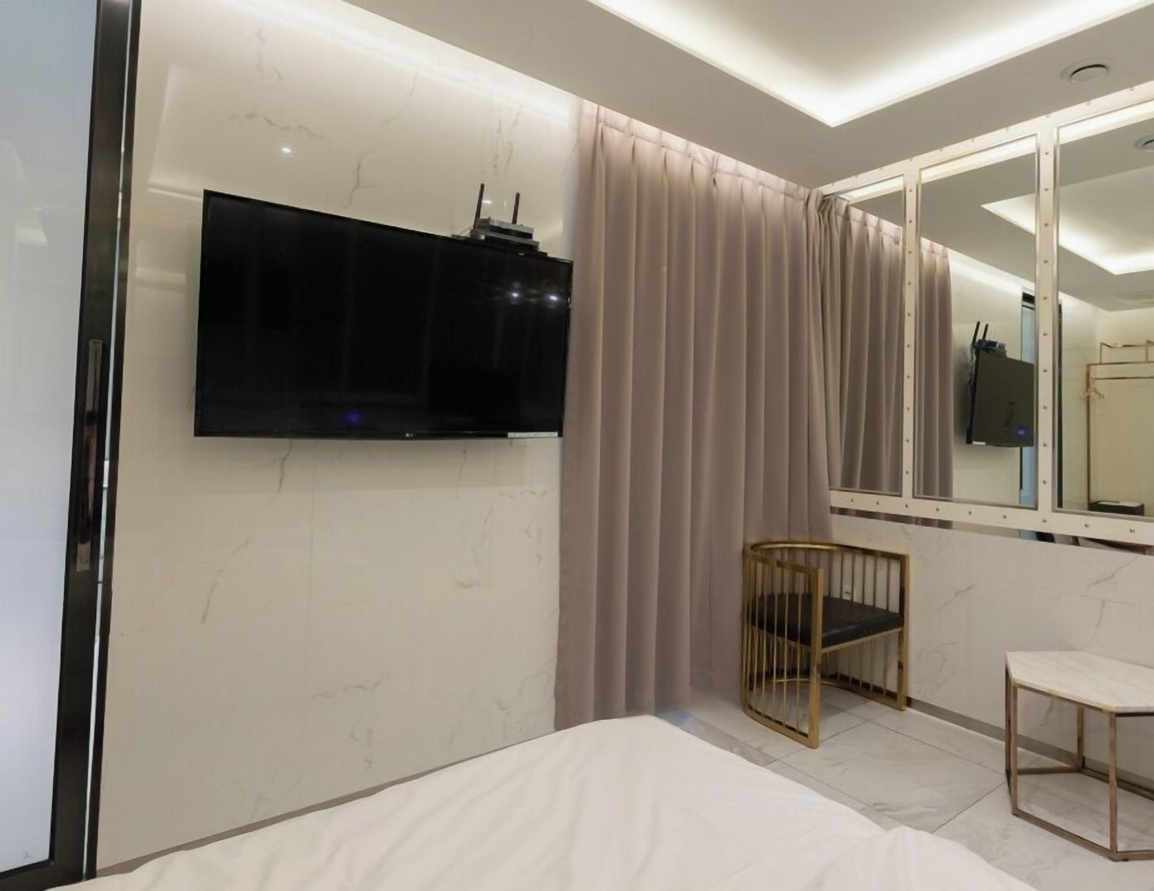 Habitación de hotel moderna con televisor de pantalla plana en la pared, cama con ropa blanca, una pequeña silla de madera y un espejo grande que refleja la habitación.