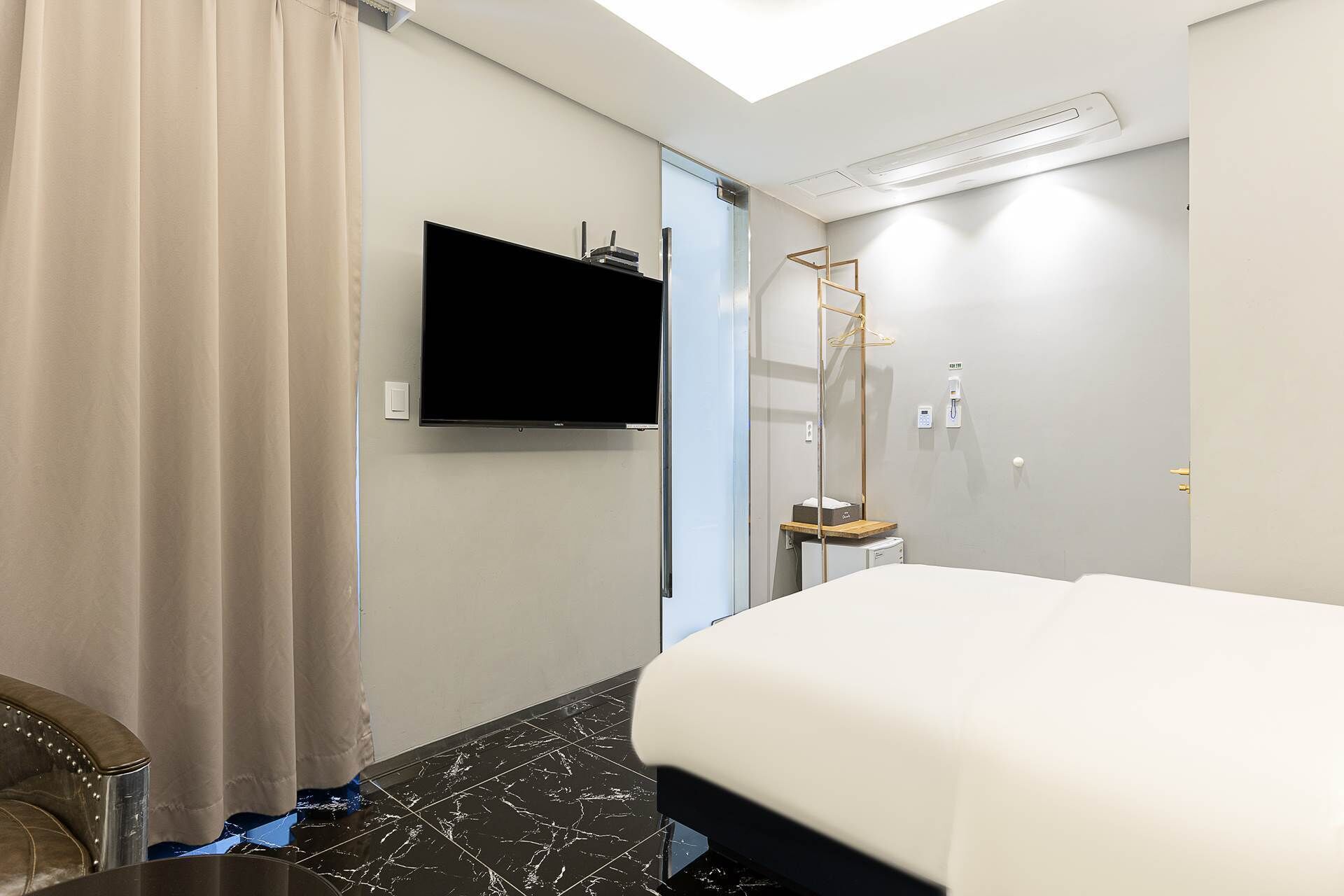 A hotel room with a bed, a flat-screen TV mounted on the wall, a shower area with a glass door, and a ceiling-mounted air conditioning unit.