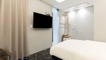 A hotel room with a bed, a flat-screen TV mounted on the wall, a shower area with a glass door, and a ceiling-mounted air conditioning unit.