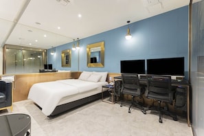 Una habitación de hotel moderna con una cama grande, un escritorio con dos computadoras y un espejo en la pared.