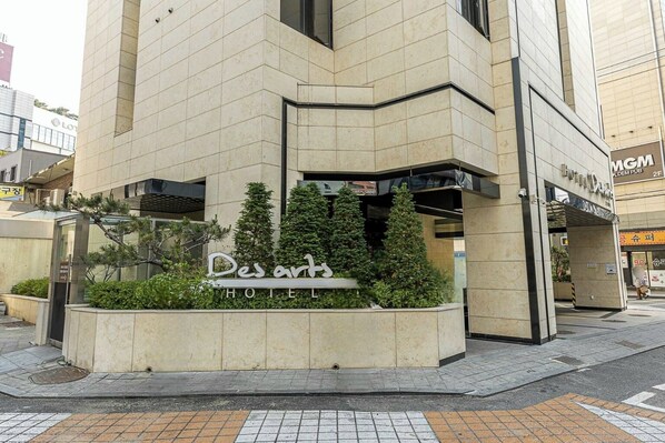 Entrada de hotel con un cartel que dice "Des arts HOTEL" y un pequeño jardín con plantas en macetas.