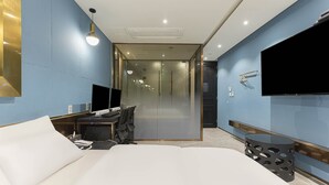 A modern hotel room with a large bed, a desk with a computer, a flat-screen TV, and a glass partition leading to a hallway. - Hotel Des Arts (Seoul)