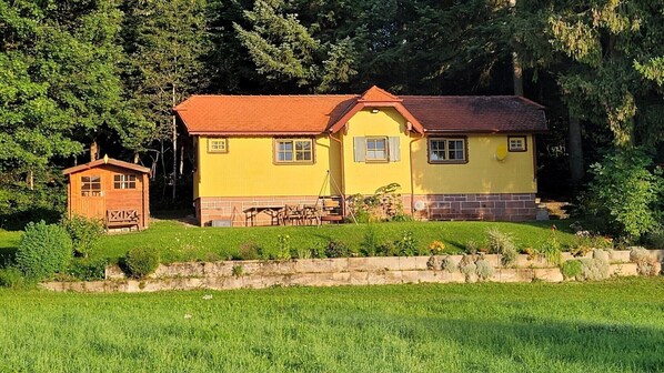 A yellow house with a red roof, a small wooden structure, and a stone wall in front.