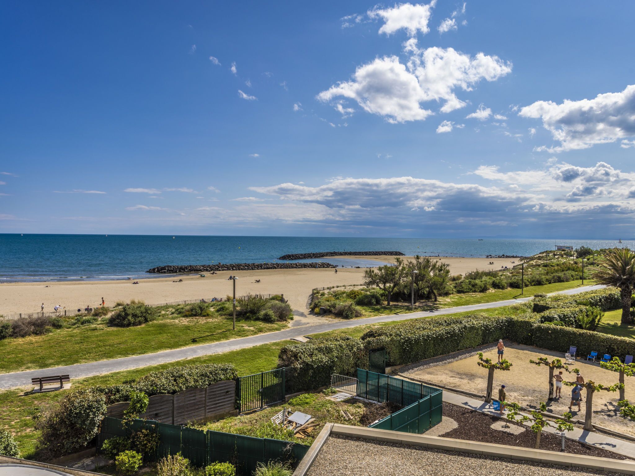A beach view with a clear sky, a sandy beach, and a grassy area with a fence and trees.