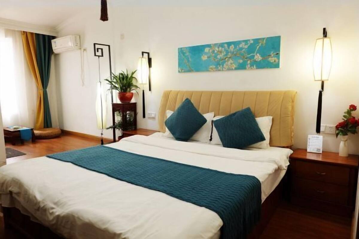 A hotel room with a bed, a potted plant, a painting, and a nightstand.