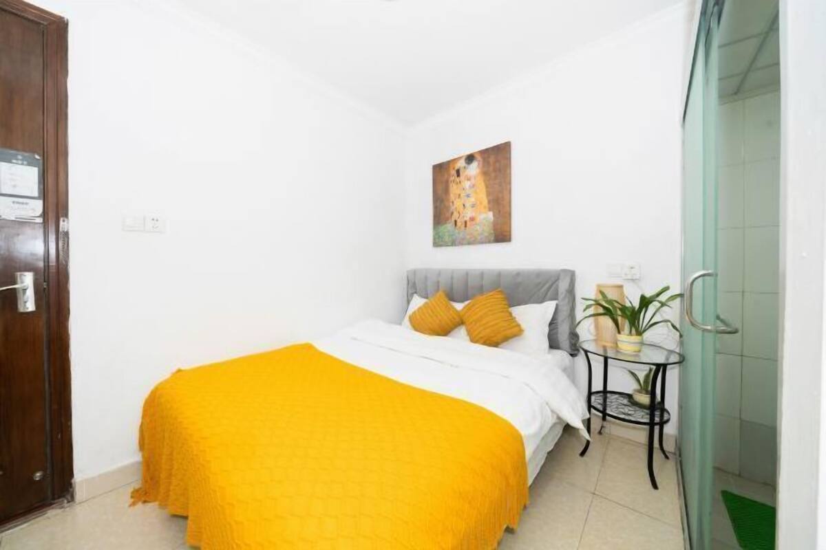 A bedroom with a bed, a painting, a small table with a plant, and a door.