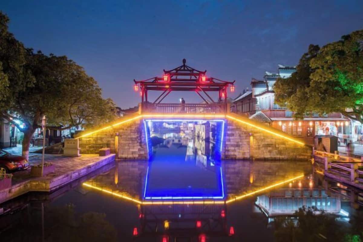 A traditional bridge with a lantern-lit walkway over water, surrounded by trees and buildings.