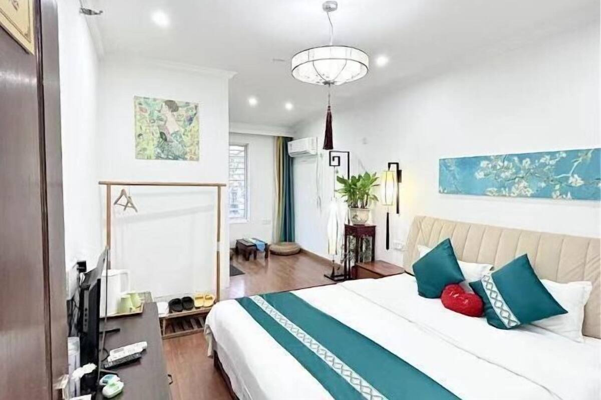 A hotel room with a bed, a desk, a chair, a plant, and a painting on the wall.