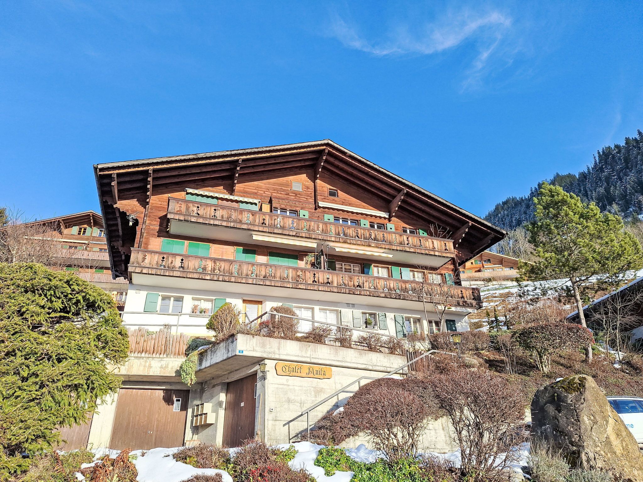 Ein traditionelles Holzhäuschen mit mehreren Balkonen, einem Schild mit der Aufschrift "Chalet Maier" und Blick auf einen verschneiten Berg.