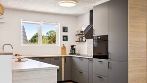 Una cocina moderna con armarios grises, encimera blanca, horno empotrado y ventana con vistas a árboles.