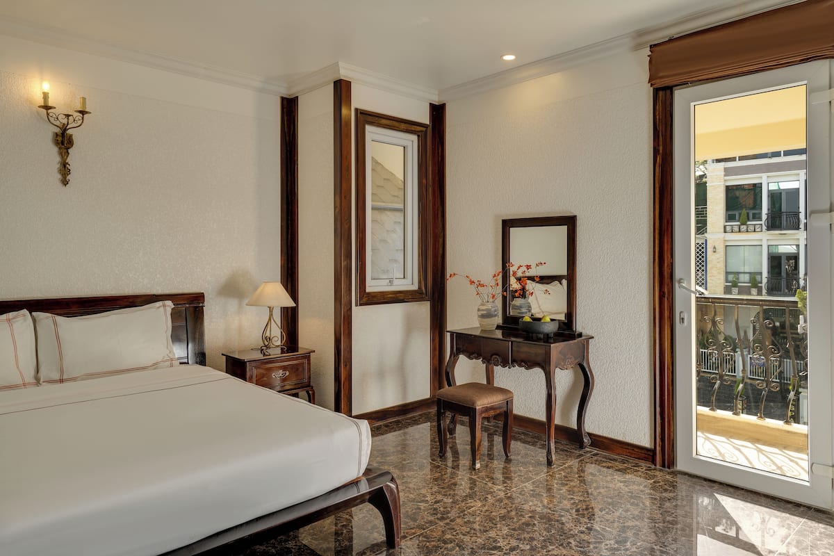 Habitación de hotel con cama, mesita de noche, lámpara, taburete pequeño de madera y un escritorio con espejo y florero.
