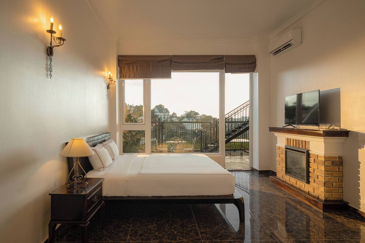 Habitación de hotel con una cama grande, televisor, chimenea y balcón con vistas al exterior.