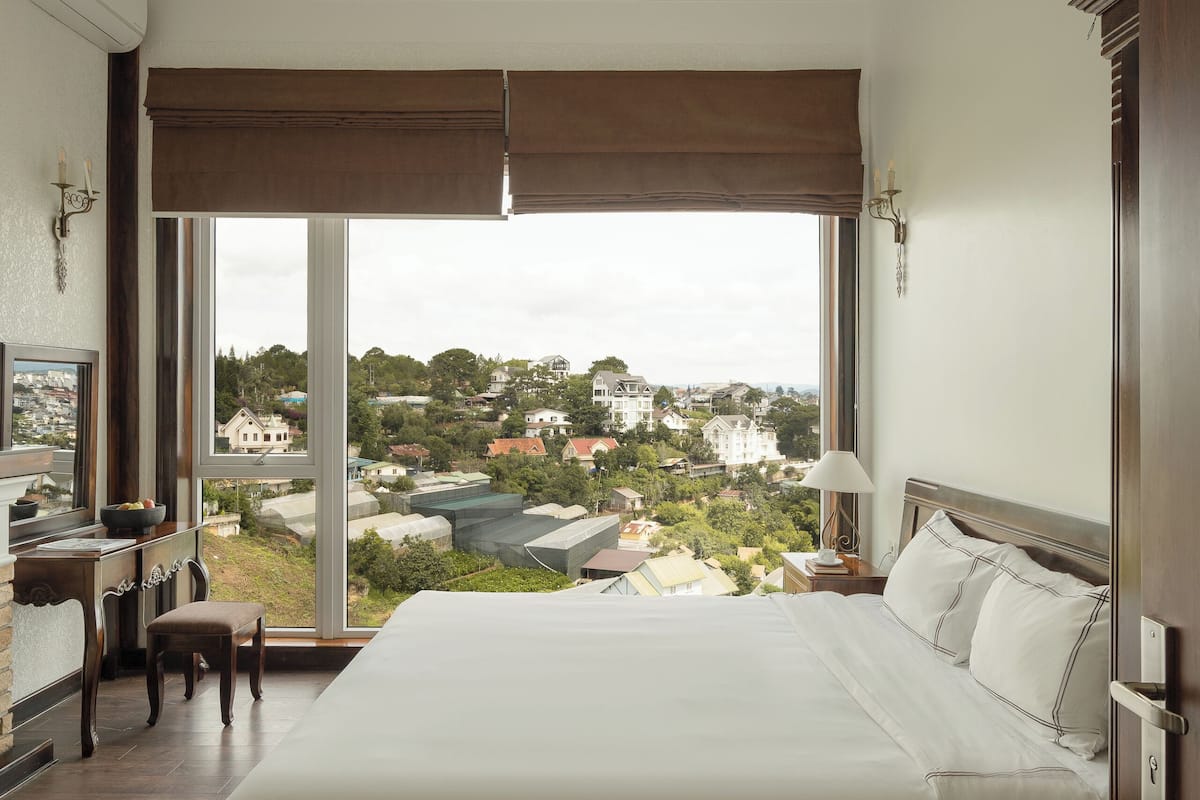 Un dormitorio con una cama grande, un escritorio y una silla, con vista a un paisaje urbano a través de amplias ventanas.