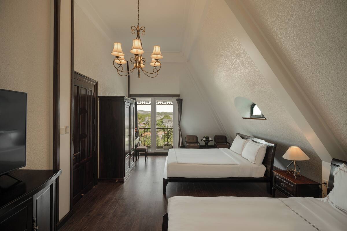 Habitación de hotel con dos camas, un candelabro, televisión y balcón con vistas.