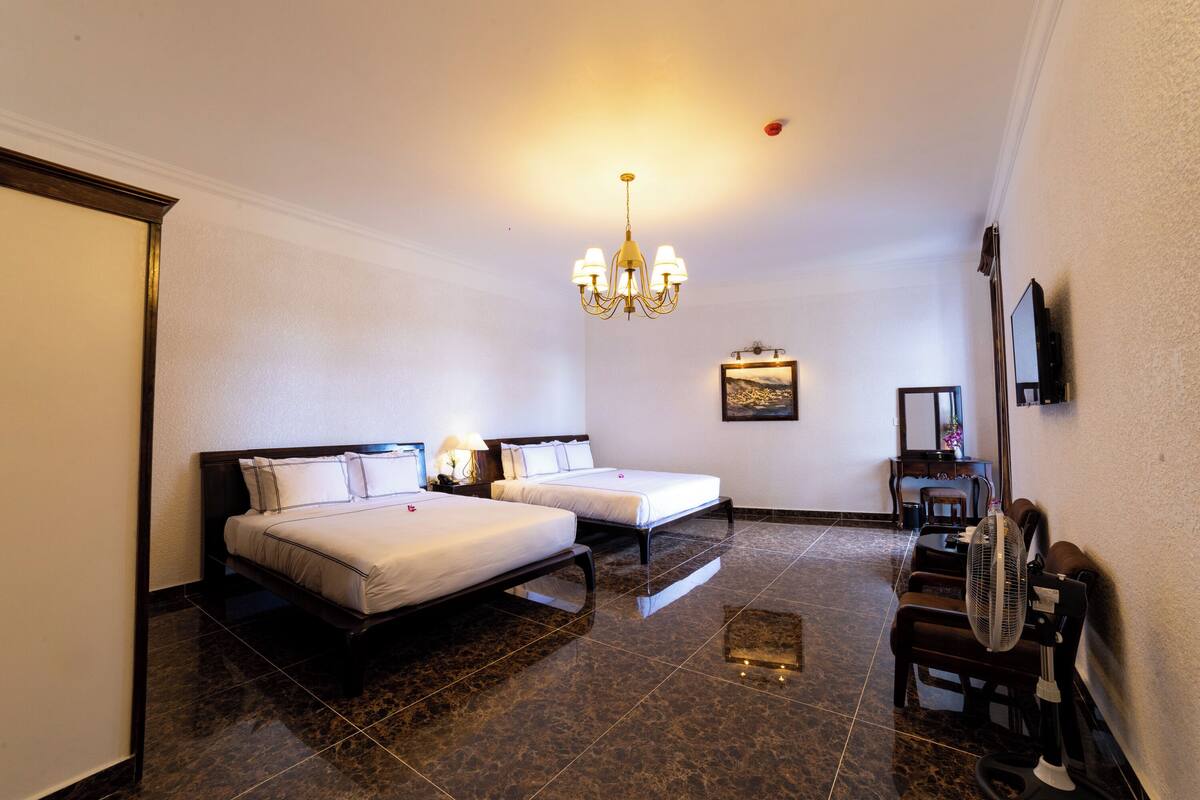Habitación de hotel con dos camas, un candelabro, un televisor de pantalla plana y un ventilador de pie.