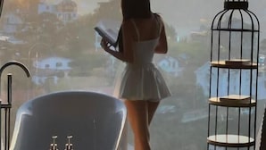 Une femme vêtue d’une robe blanche se tient près d’une grande fenêtre, d’où l’on peut admirer des maisons et un coucher de soleil.