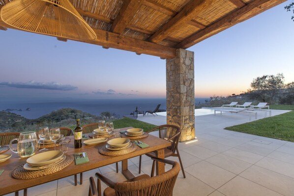 A dining table set for a meal with a view of the sea, outdoor seating, and a stone pillar.
