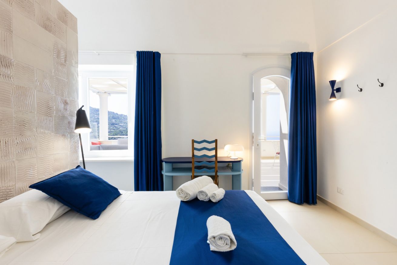A hotel room with a bed, a desk, and a chair. There are curtains, a lamp, and a view of the outside.