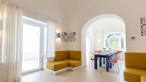 A modern living room with a sofa, dining table, and chairs. There is a large window and a door leading to an outdoor area.