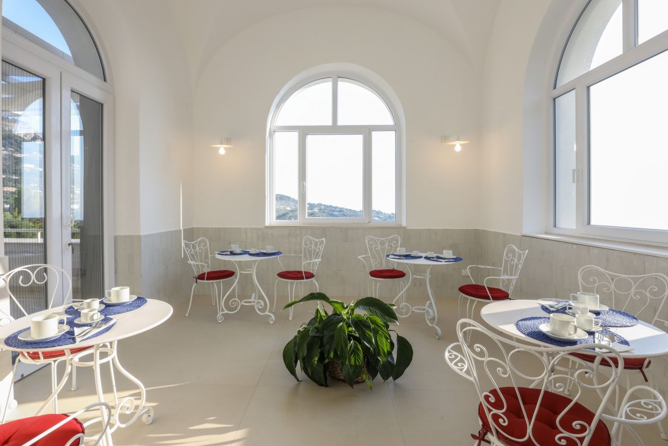A sunlit room with white metal chairs and tables, a large arched window, and a potted plant in the center.