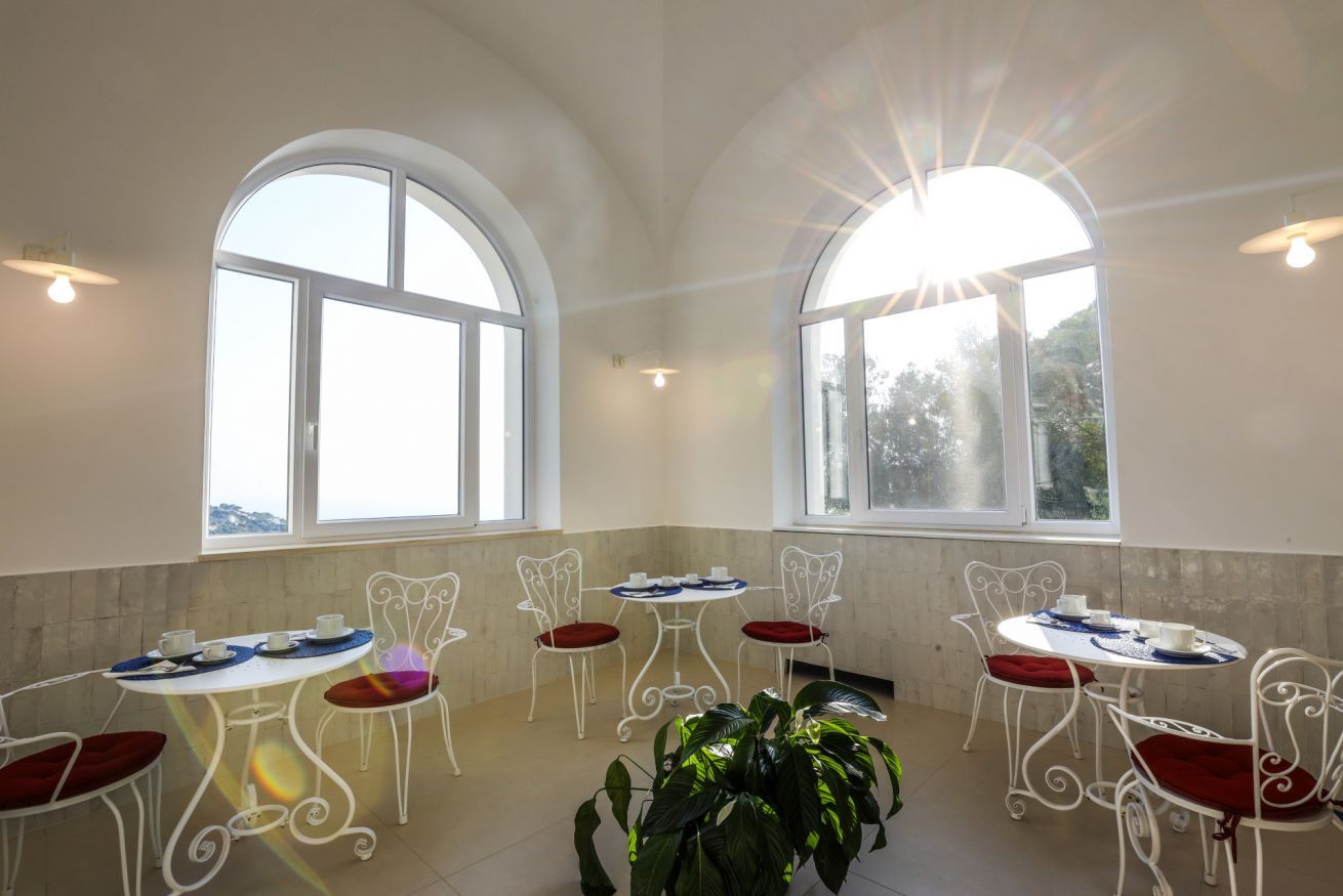A dining area with white metal chairs and tables, large arched windows, and a potted plant.