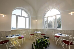 A dining area with white metal chairs and tables, large arched windows, and a potted plant.