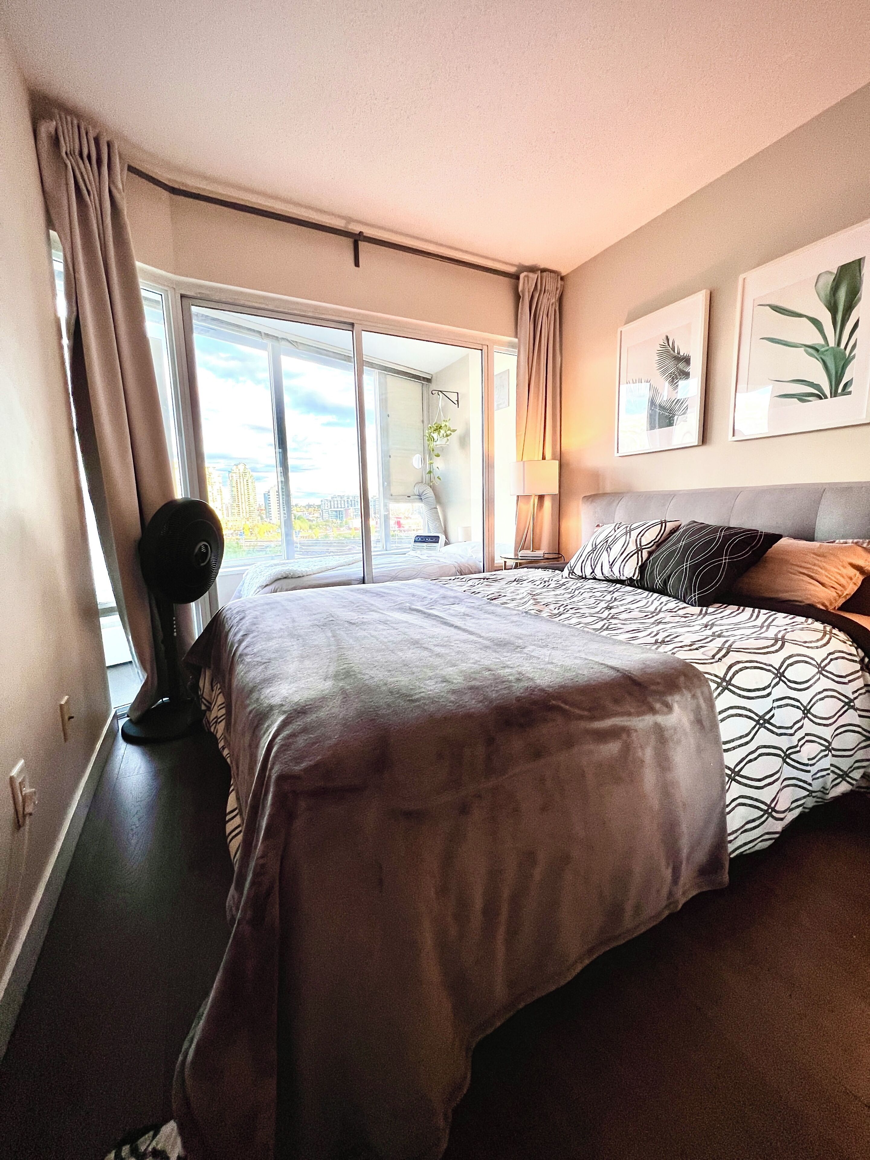 Habitación de hotel con una cama grande, un ventilador de pie, vistas de la ciudad a través de un ventanal amplio y dos cuadros en la pared.
