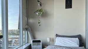 A hotel room with a large window, a bed with a white textured blanket, a bedside table with a lamp, and a wall-mounted air purifier.