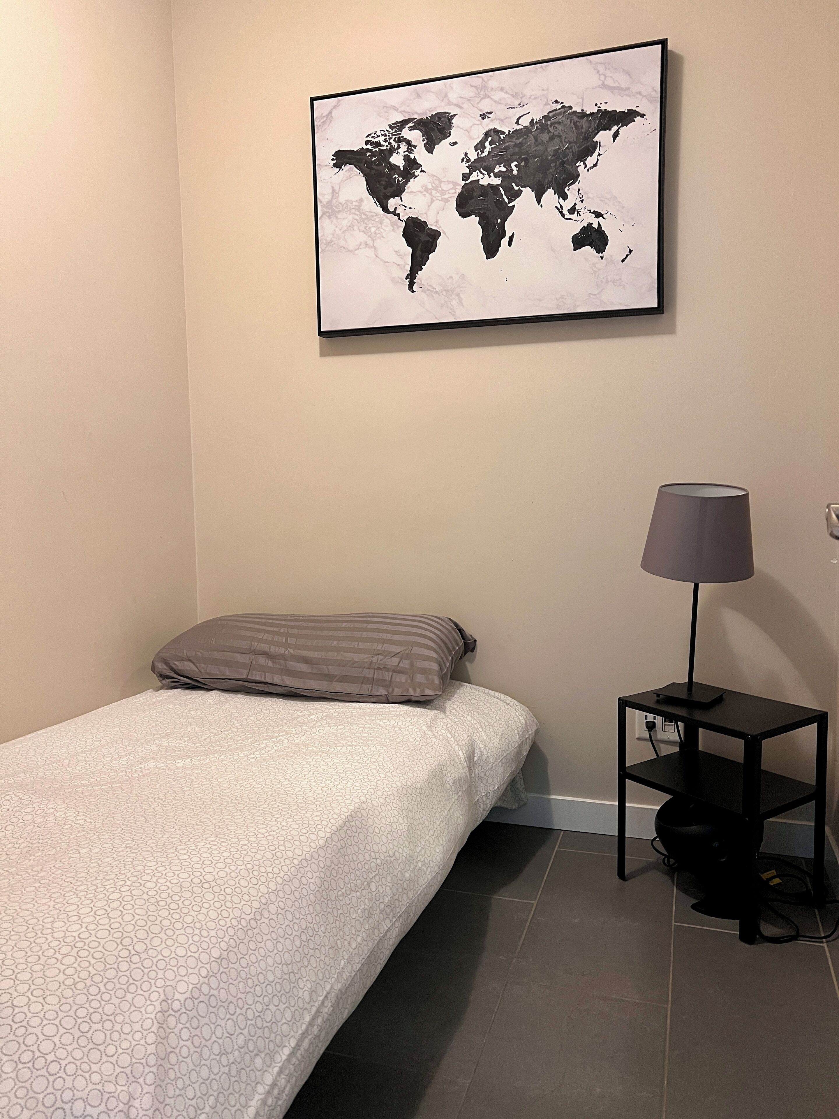 Una cama individual con una colcha estampada, una almohada gris, una obra de arte con un mapa del mundo en la pared, una lámpara sobre una mesita de noche y un enchufe en la pared.