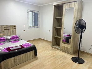Ein Schlafzimmer mit einem Bett, einem Holzschrank, einem Standventilator und einem Fenster.