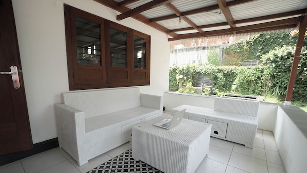 A white wicker sofa and ottoman set in a covered patio area with a tiled floor and a geometric patterned rug.