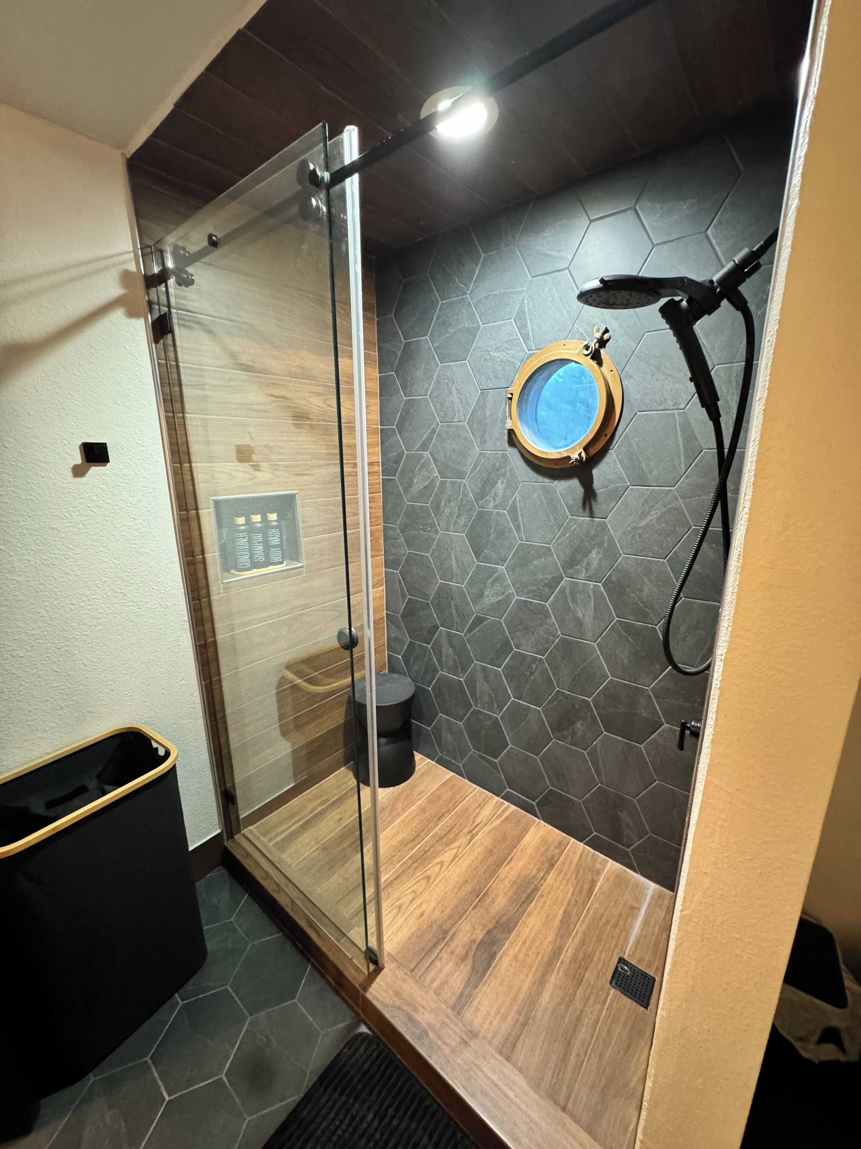 A modern shower area with a glass door, a wooden floor, and a hexagonal tile wall.