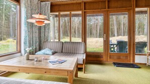 L’intérieur chaleureux d’un chalet avec un plafond en bois, un canapé agrémenté de coussins, une table à manger et de grandes fenêtres offrant une vue sur la forêt.