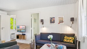 Un salon moderne avec un canapé gris, une table basse et un téléviseur. Des lampes murales et des tableaux encadrés ornent le mur.