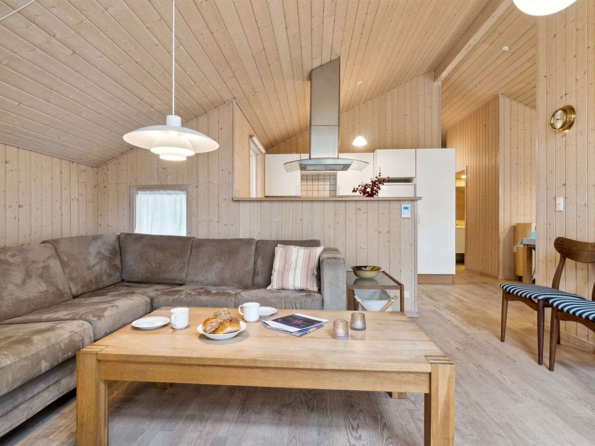 A cozy living room with a wooden ceiling, a sofa, a coffee table with pastries, and a dining area with a chair.