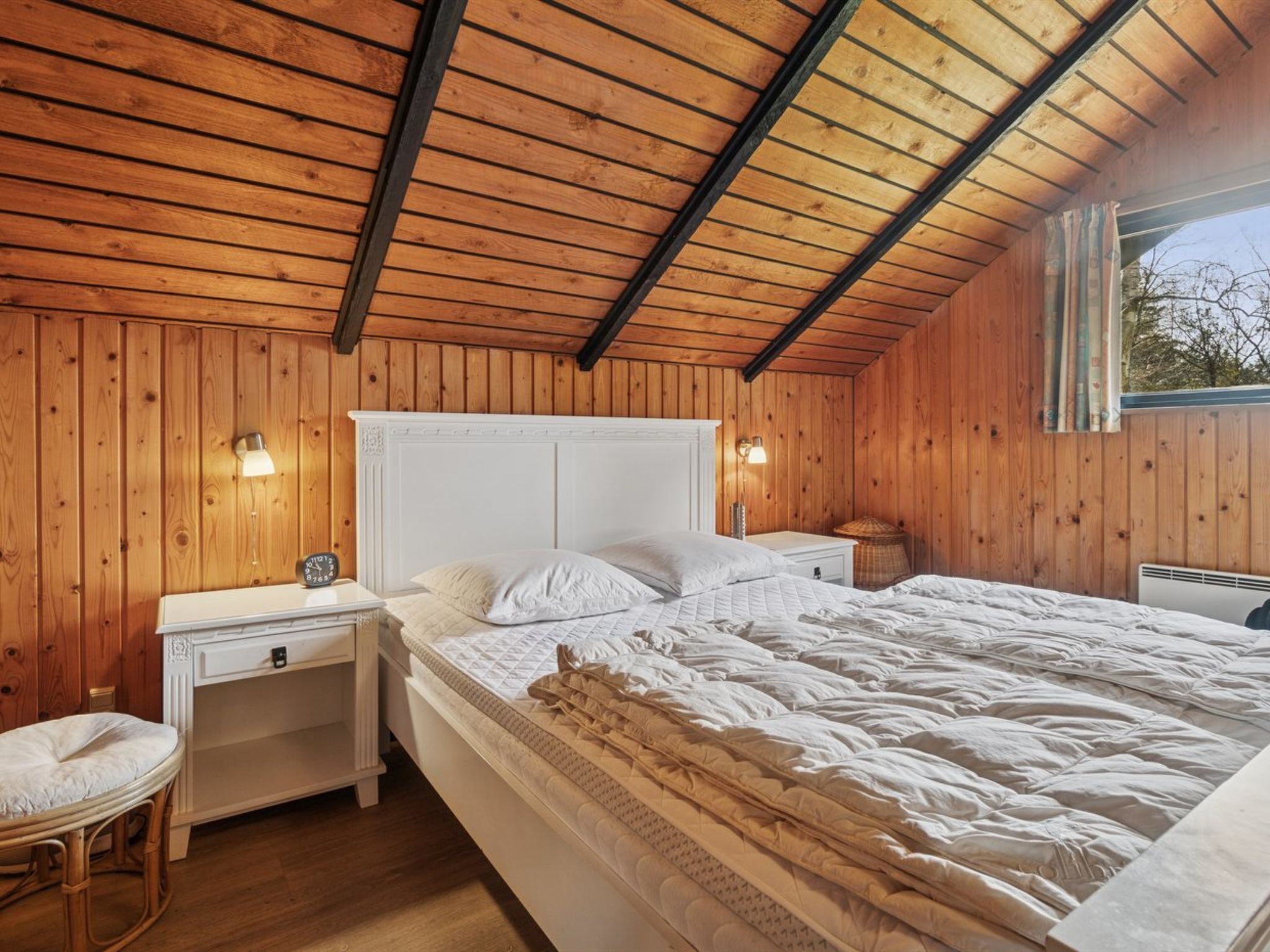 A cozy bedroom with a wooden ceiling, a large bed, bedside tables, and a window.