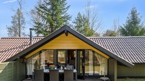 Un espace de restauration extérieur moderne, doté d’une terrasse en bois et d’une structure vitrée, le tout entouré d’arbres.