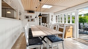 Un espace repas avec une longue table en bois, des chaises blanches et des sièges noirs rembourrés. Des luminaires suspendus sont placés au-dessus de la table, et une grande fenêtre laisse entrer la lumière naturelle.