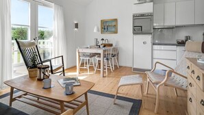 A bright, open-plan living and dining area with a wooden dining table, white chairs, a kitchen with white cabinets, and a large window with curtains.
