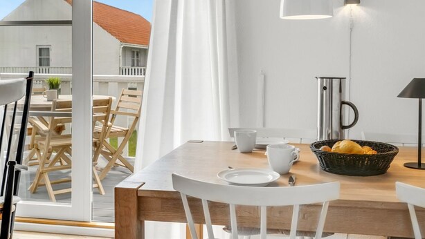 A dining area with a wooden table, white chairs, a basket of bread, a coffee pot, and a candle on the table.