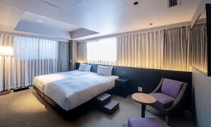 A modern hotel room with a large bed, a small desk, a chair, and a sofa.
