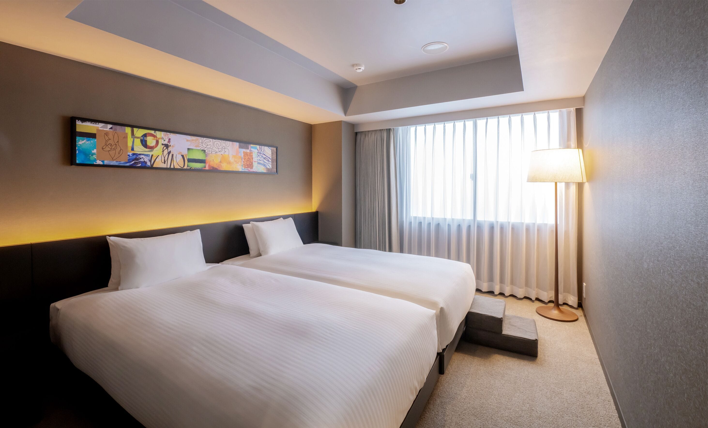 A hotel room with two beds, a ceiling light, a painting on the wall, and a window with curtains.