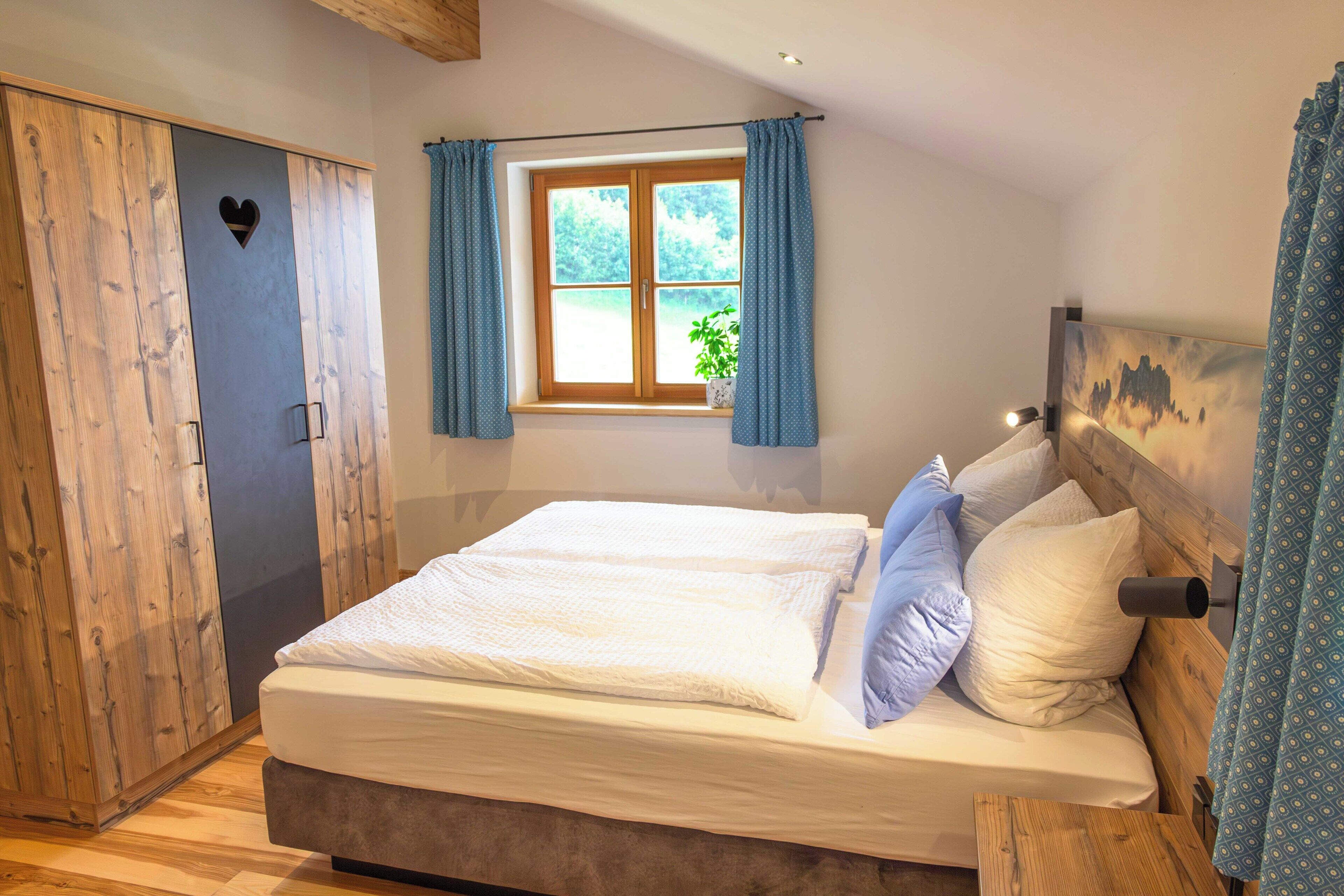 A bedroom with a bed, wooden wardrobe, a window with curtains, and a view of greenery outside.