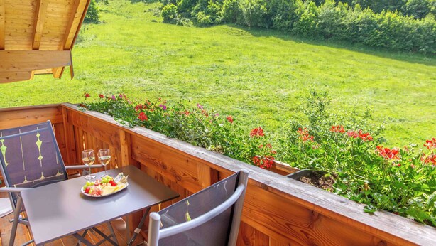 A wooden deck with a table set for two, overlooking a grassy area with flowers and trees. - Ferienwohnung Kampenwand (Bernau am Chiemsee)