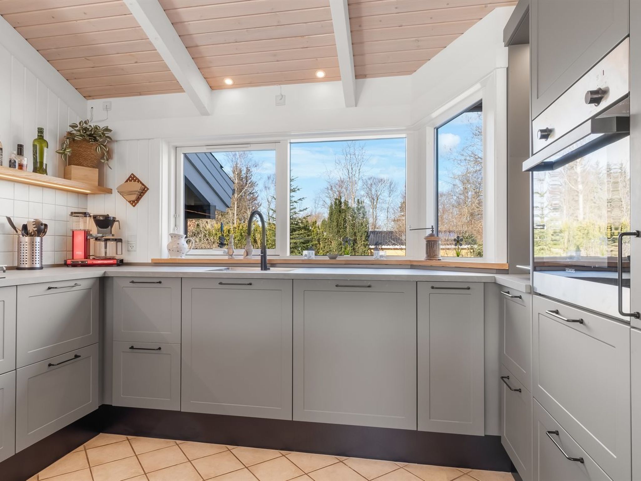 A modern kitchen with grey cabinets, a large window, and a wooden ceiling.