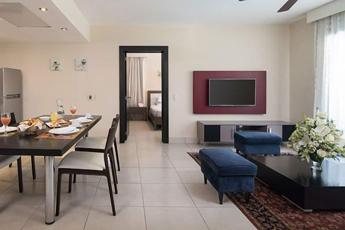 Una cocina moderna con comedor, una sala de estar con televisor, y una zona de estar con mesa de centro y flores.