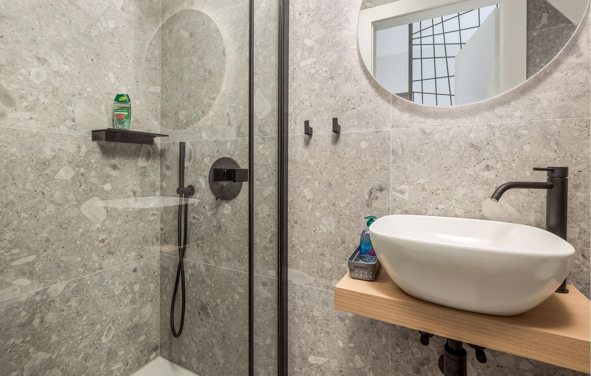 A bathroom with a white sink, a handheld showerhead, a small shelf with a bottle, and a window with a grid pattern.
