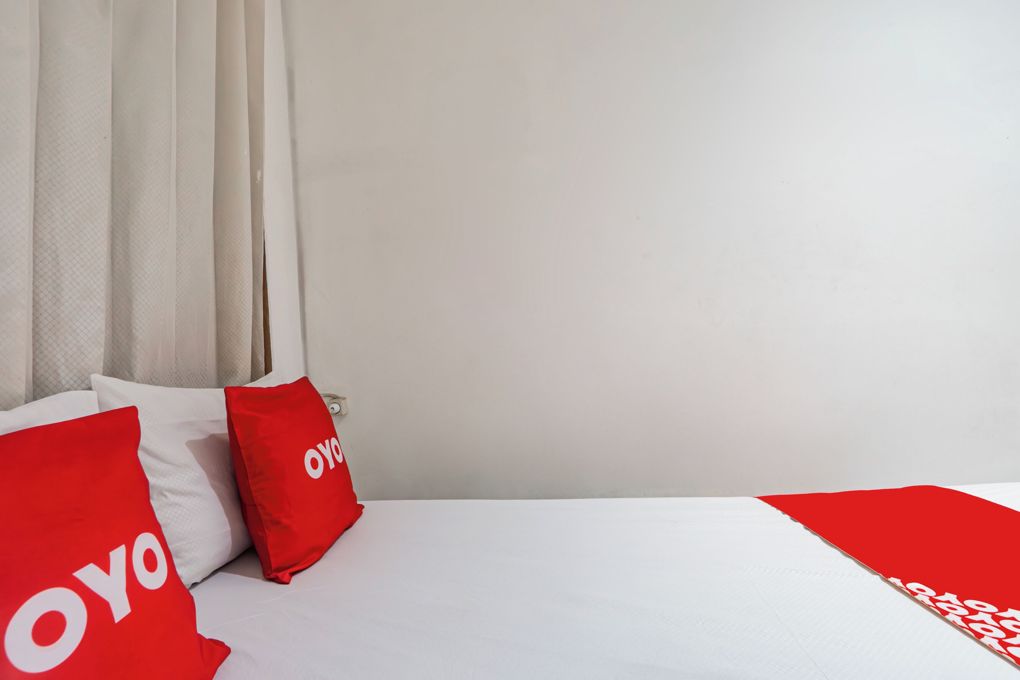 A bed with white bedding and red pillows with "YOYO" text, a white curtain, and a plain wall.